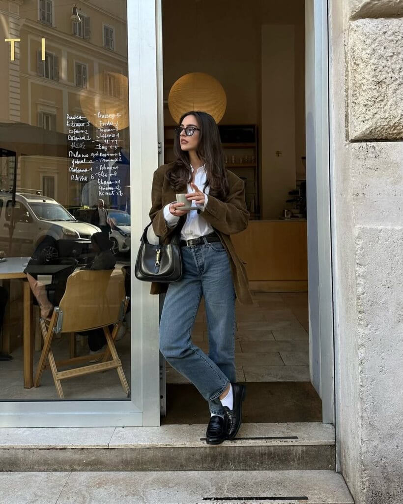 Woman in brown blazer, white shirt, blue jeans, black loafers, standing with coffee; loafers outfit, casual chic.