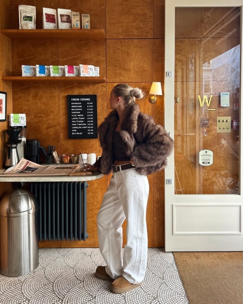 Woman in faux fur jacket, brown top, white pants, and Ugg boots at coffee bar; chic winter Ugg outfit.