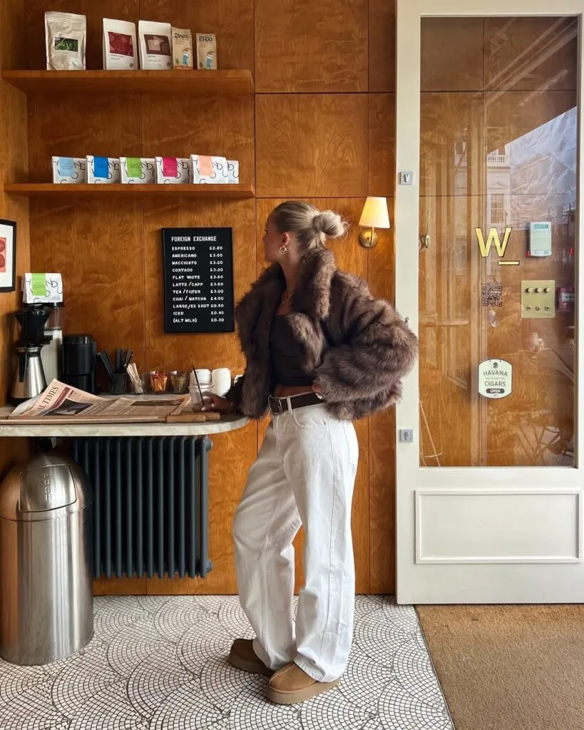 Woman in faux fur jacket, brown top, white pants, and Ugg boots at coffee bar; chic winter Ugg outfit.
