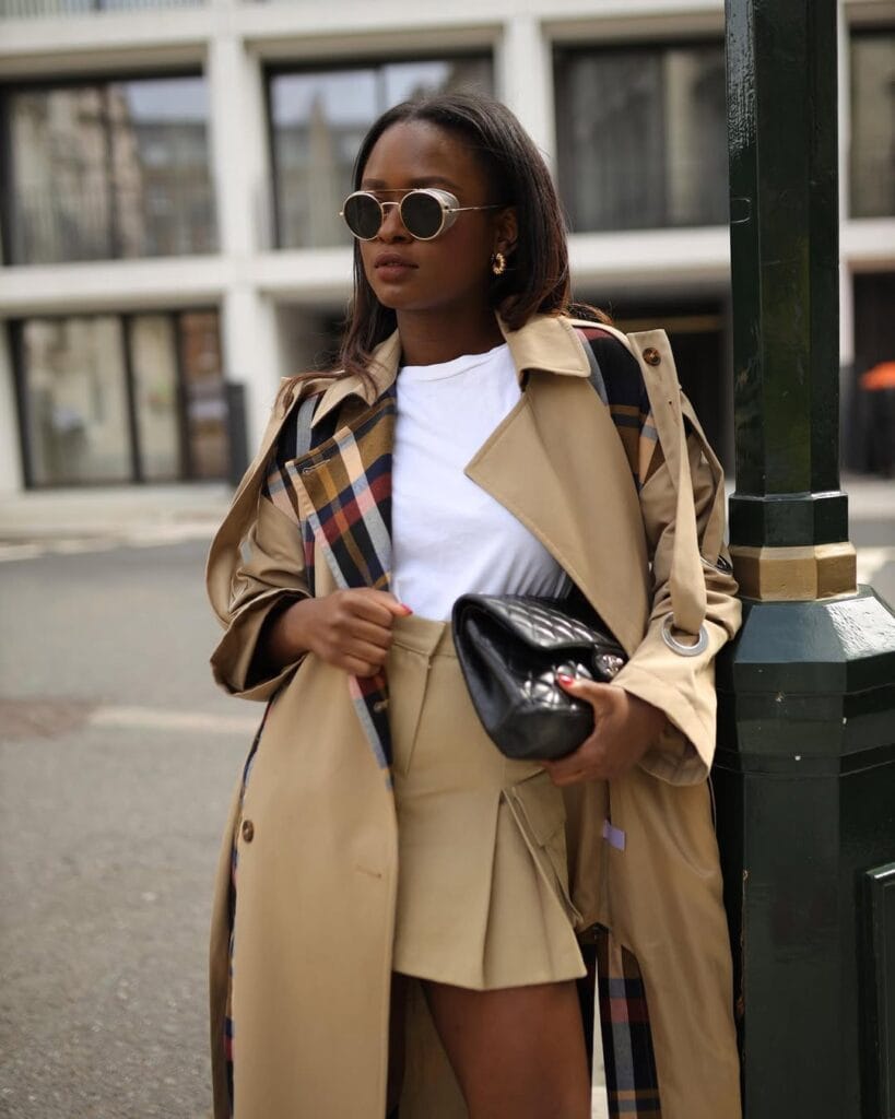 Woman in trench coat with checked lining, khaki mini skirt, white t-shirt, round sunglasses, and black bag