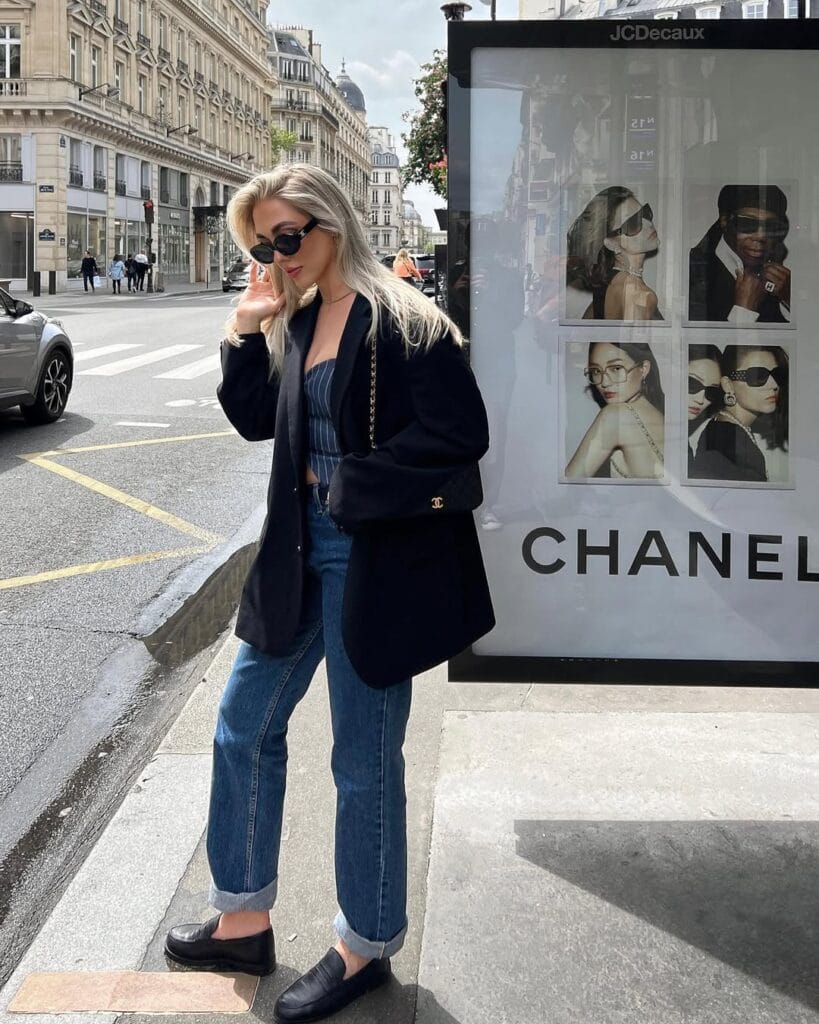 Blonde woman in black blazer, blue jeans, black loafers, sunglasses, and chain bag; Parisian loafers outfit