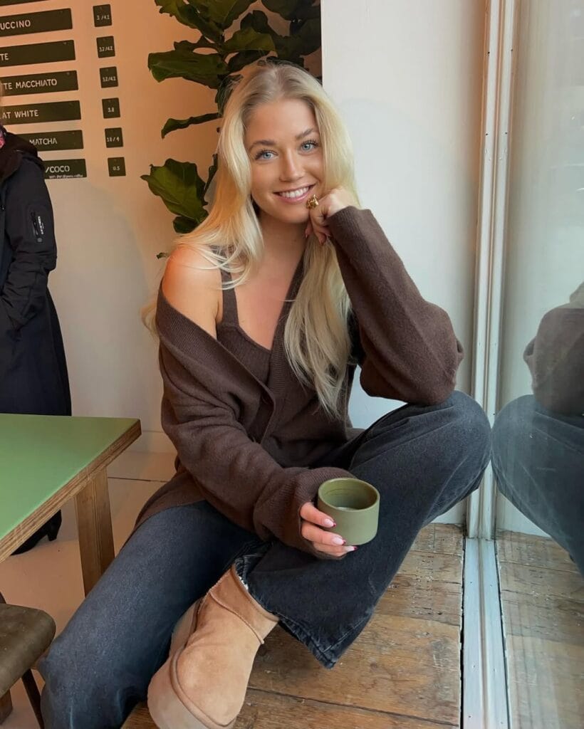 Woman in brown knit set, charcoal jeans, and Ugg boots by window with green cup; relaxed Ugg outfit.