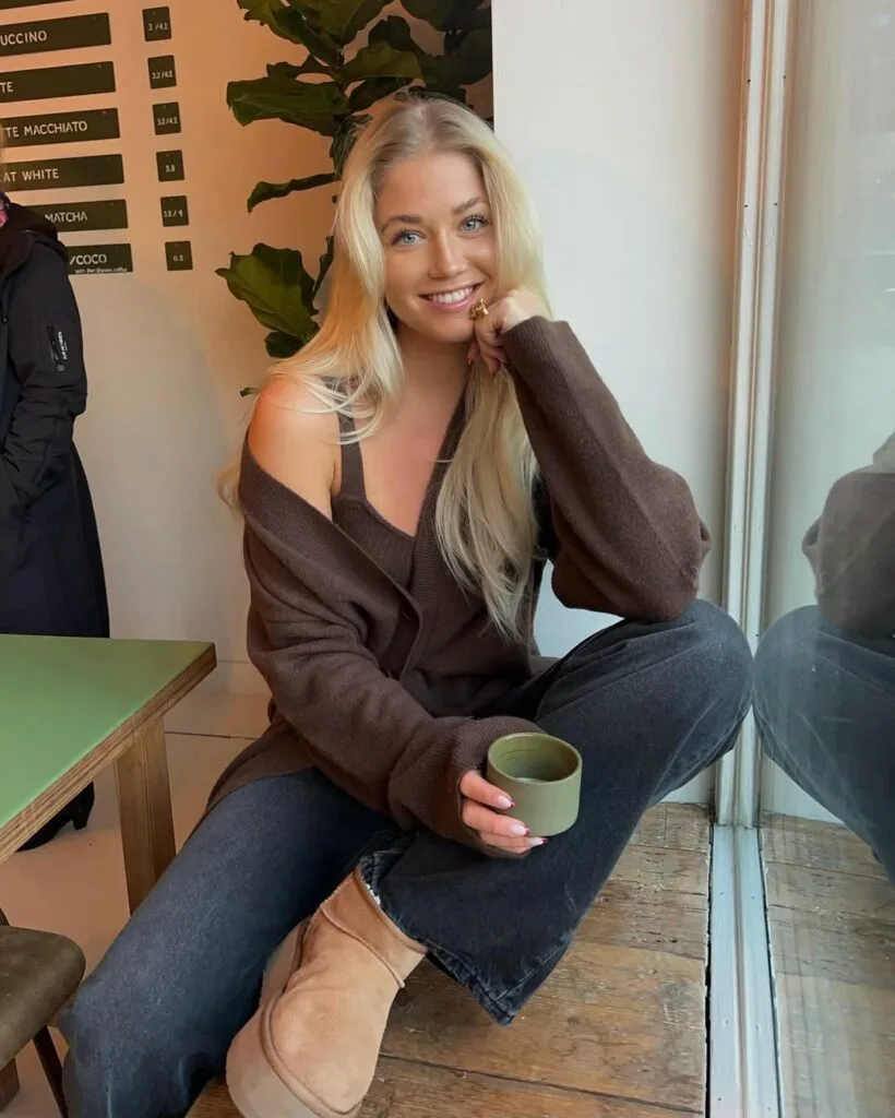 Woman in brown knit set, charcoal jeans, and Ugg boots by window with green cup; relaxed Ugg outfit.
