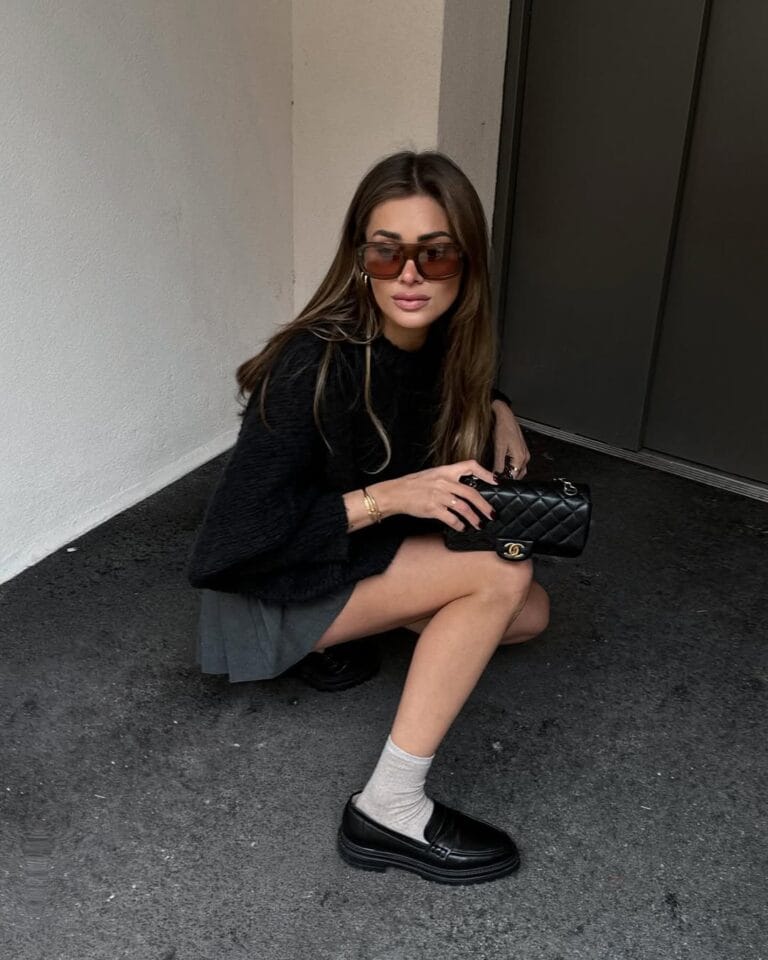 Woman in black fuzzy sweater, pleated mini, beige socks, black loafers, sunglasses, and Chanel bag; textured loafers look.