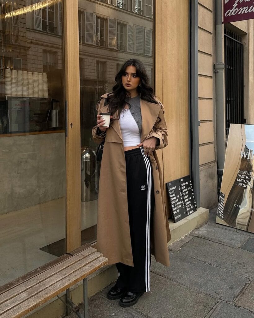 Woman in trench coat, black track pants, cropped tee, gray sweater, and black oxfords outside coffee shop