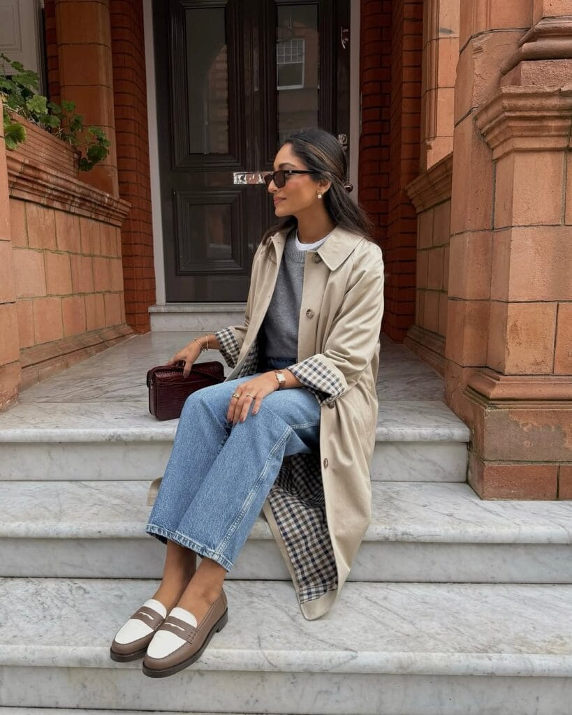 Woman in light trench coat, checkered lining, wide-leg jeans, and loafers sitting on marble steps