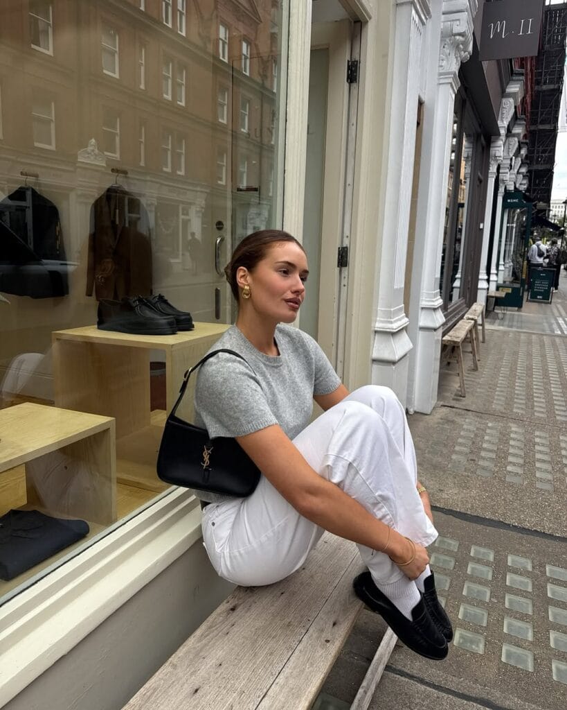 Woman in gray knit tee, white jeans, black loafers, white socks, YSL bag; clean loafers outfit on city bench.