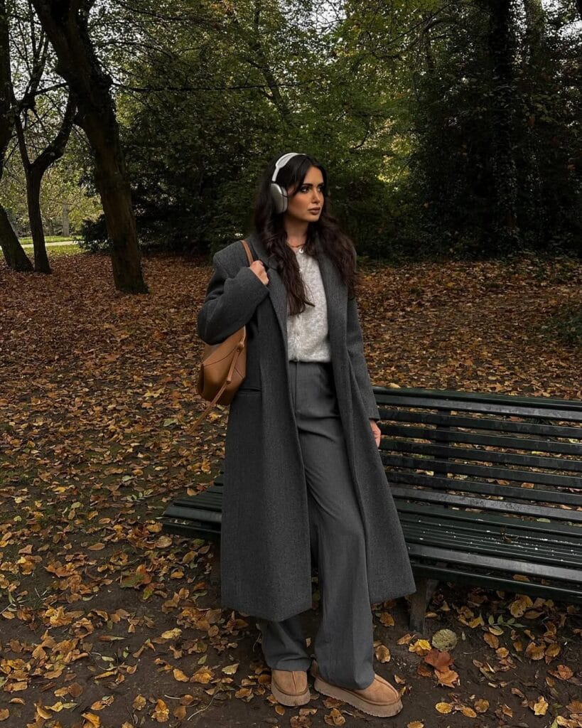Woman in long grey coat, headphones, wide pants, and Ugg boots in autumn park; chic cozy Ugg outfit.