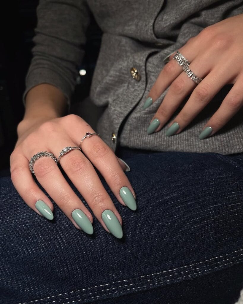 Almond-shaped old money nails in dusty sage green, styled with silver rings and grey sweater.
