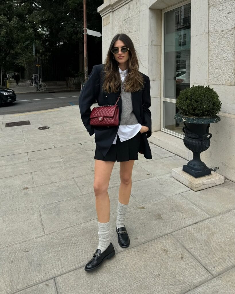 Woman in navy blazer, layered sweater, mini skirt, slouchy socks, black loafers, red Chanel bag; preppy loafers outfit.