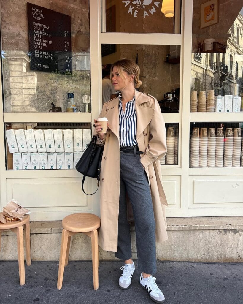Woman in trench coat, striped shirt, gray trousers, and white sneakers holding coffee outside café