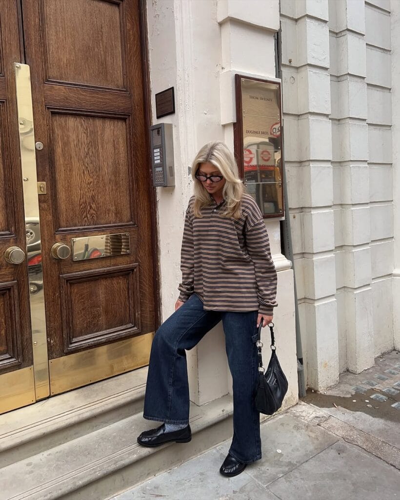 Blonde woman in striped rugby, wide-leg jeans, black loafers, chunky sunglasses, black shoulder bag; casual loafers outfit