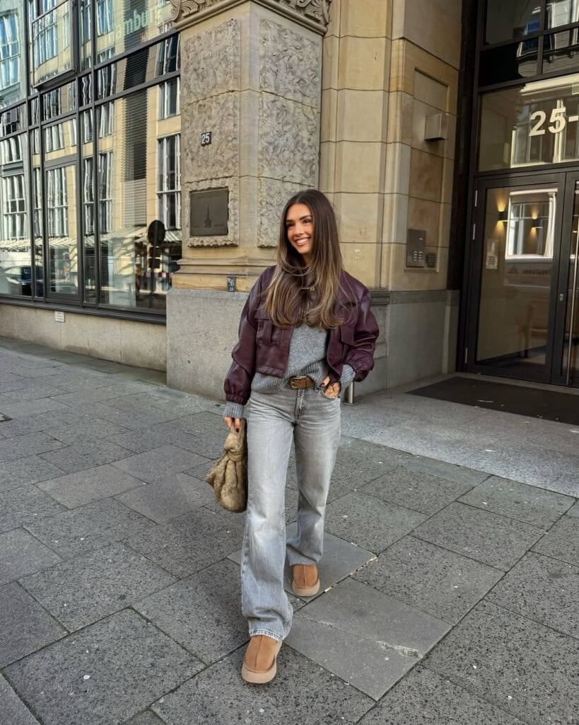 Woman in plum jacket, grey sweater, jeans, and Ugg boots, standing on city street; trendy Ugg outfit idea.