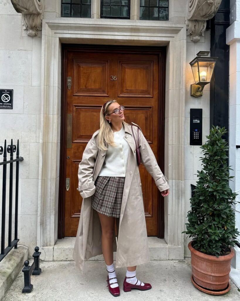 Woman in trench coat, plaid mini skirt, sweater, and burgundy Mary Janes outside brown door