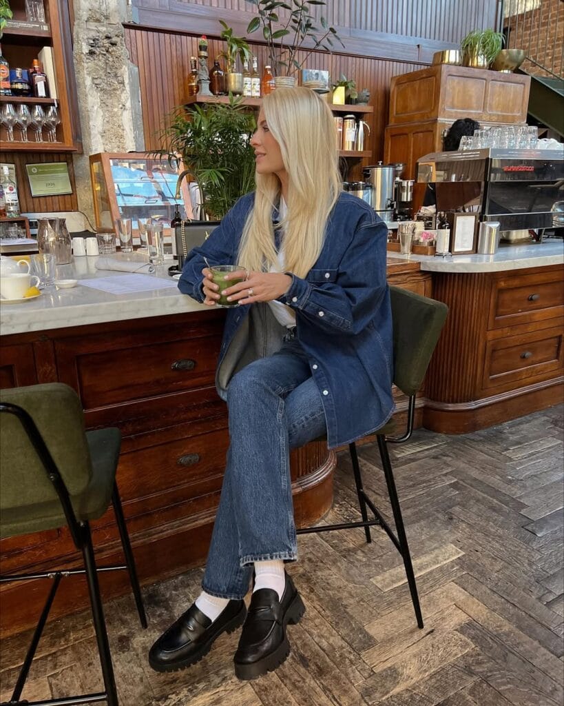 Blonde woman in blue denim jacket, jeans, white socks, black loafers, sitting at café; denim loafers outfit.