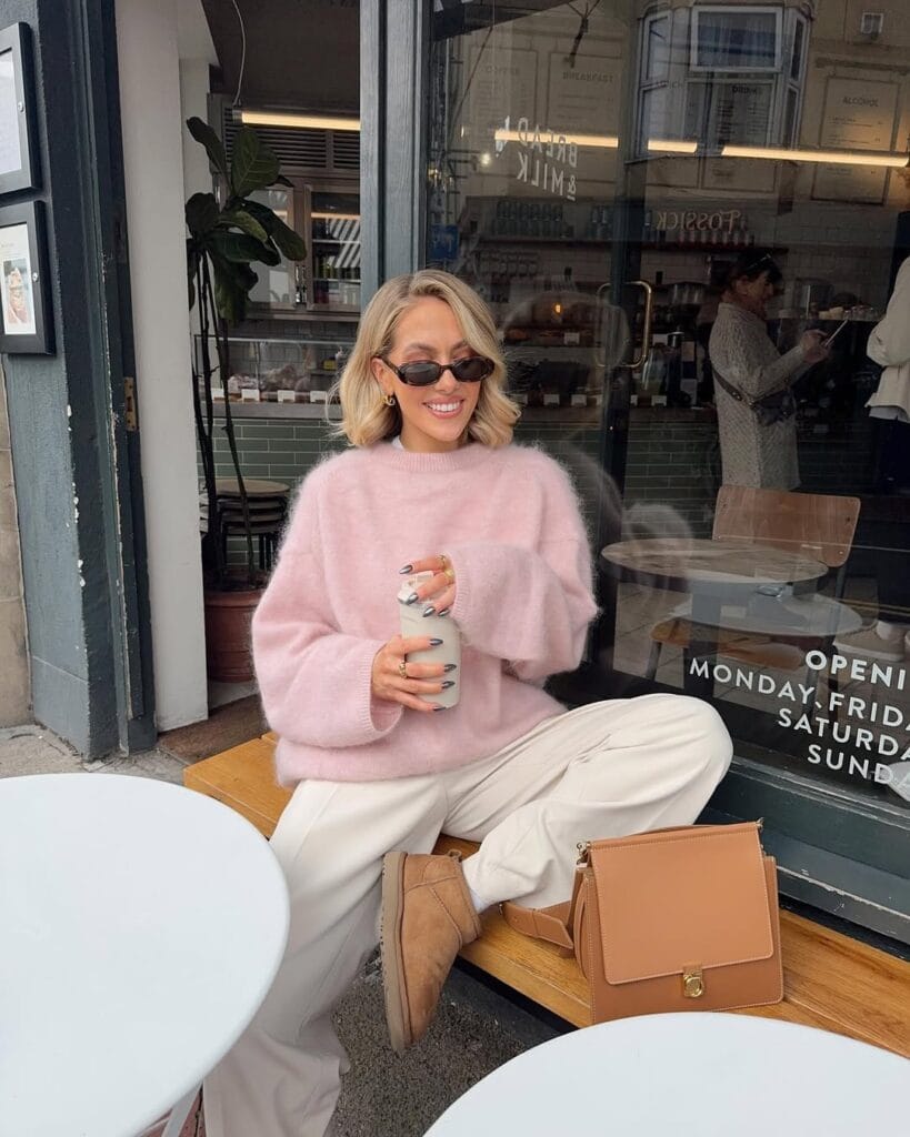 Woman in pink fuzzy sweater, white trousers, and Ugg boots with tan bag, smiling at café; cozy chic Ugg outfit.
