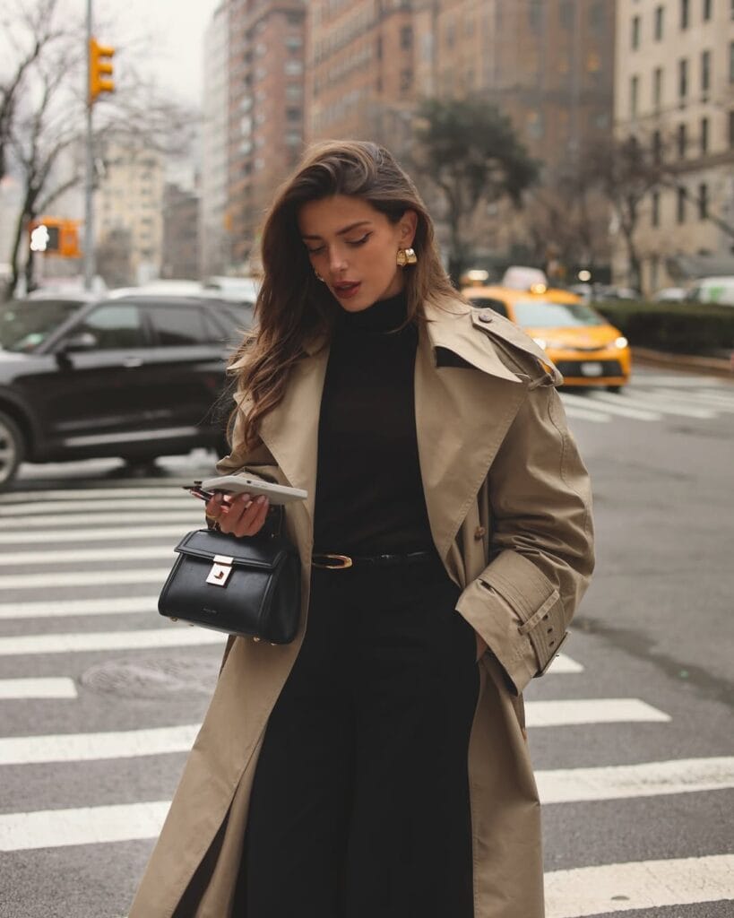 Woman in tan trench coat, black turtleneck, black pants, and gold earrings crossing city street