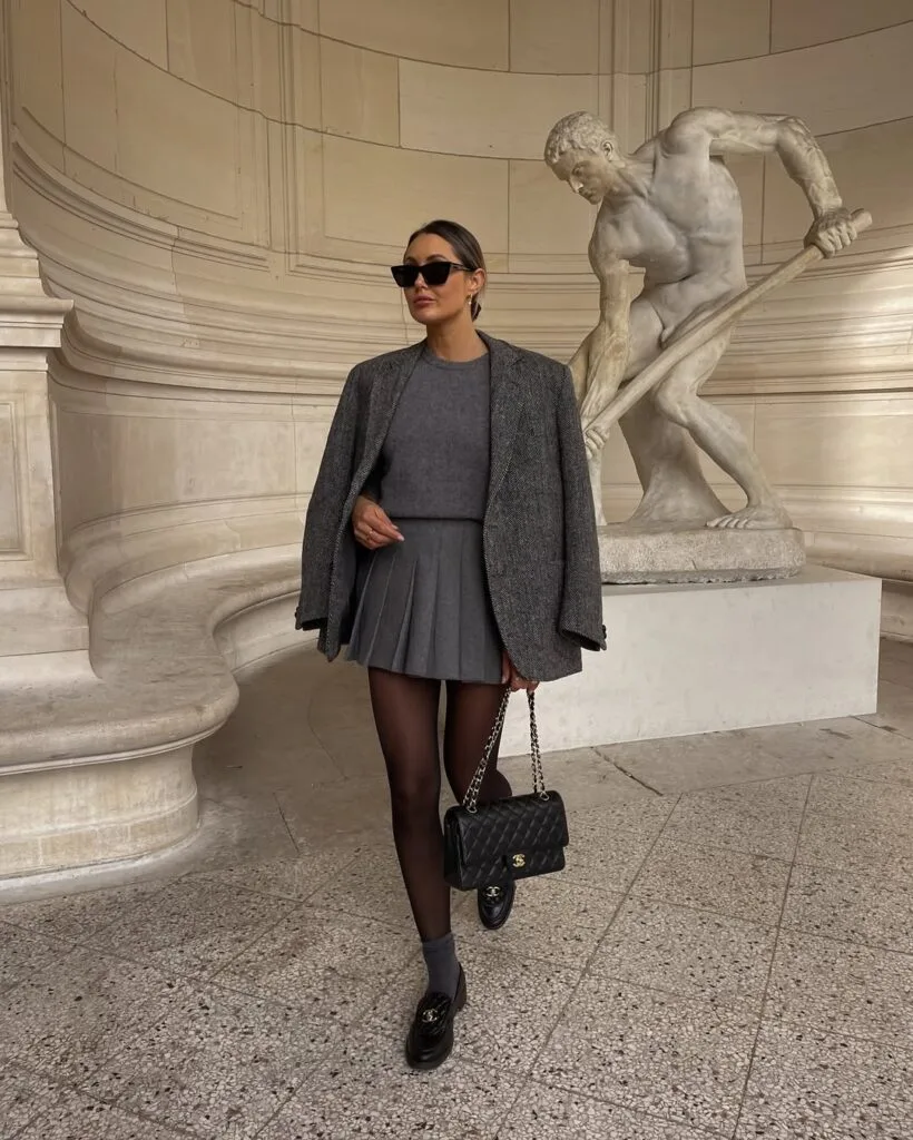 Woman in gray blazer, matching mini skirt, gray socks, Chanel loafers, sunglasses, quilted bag; luxe loafers outfit.