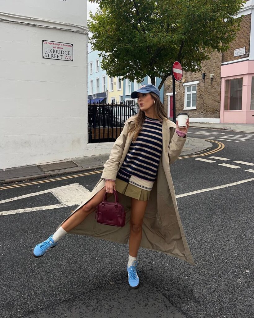 Woman in long trench coat, striped sweater, skirt, blue cap, and sneakers; sporty street style