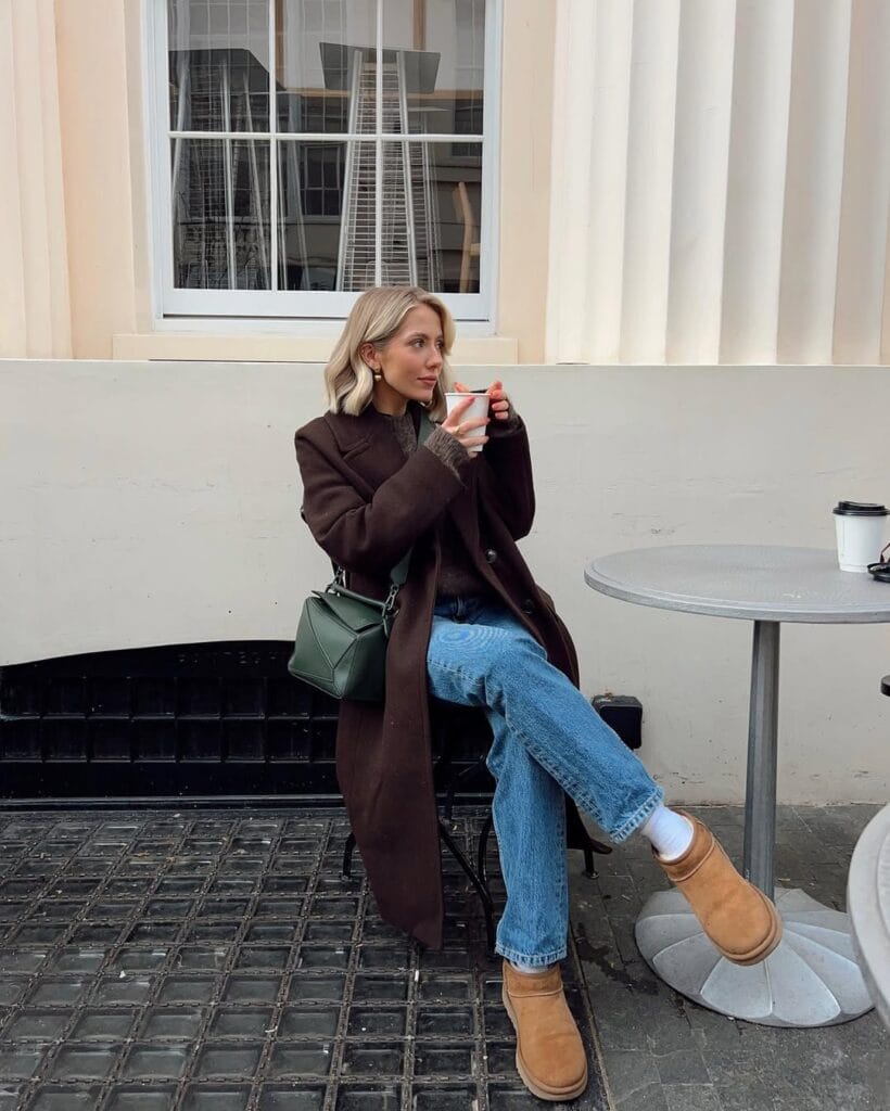 Woman wearing brown coat, jeans, and Ugg boots, sitting at café table; chic Ugg outfit idea.