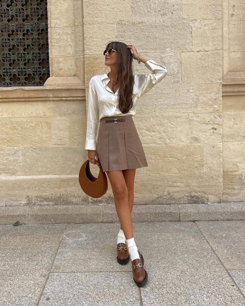 Woman in satin shirt, taupe mini skirt, white socks, brown loafers, holding round bag; elegant loafers outfit outside.