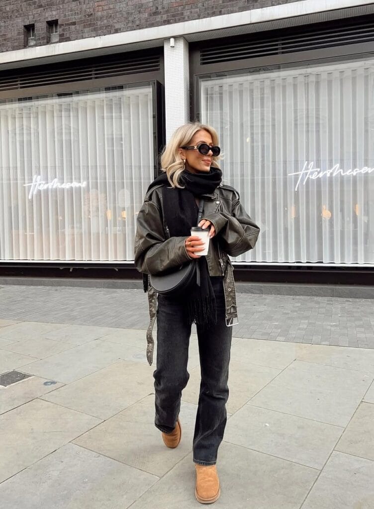 Woman in black leather jacket, scarf, dark jeans, and Ugg boots walking in city; edgy Ugg outfit for fall.
