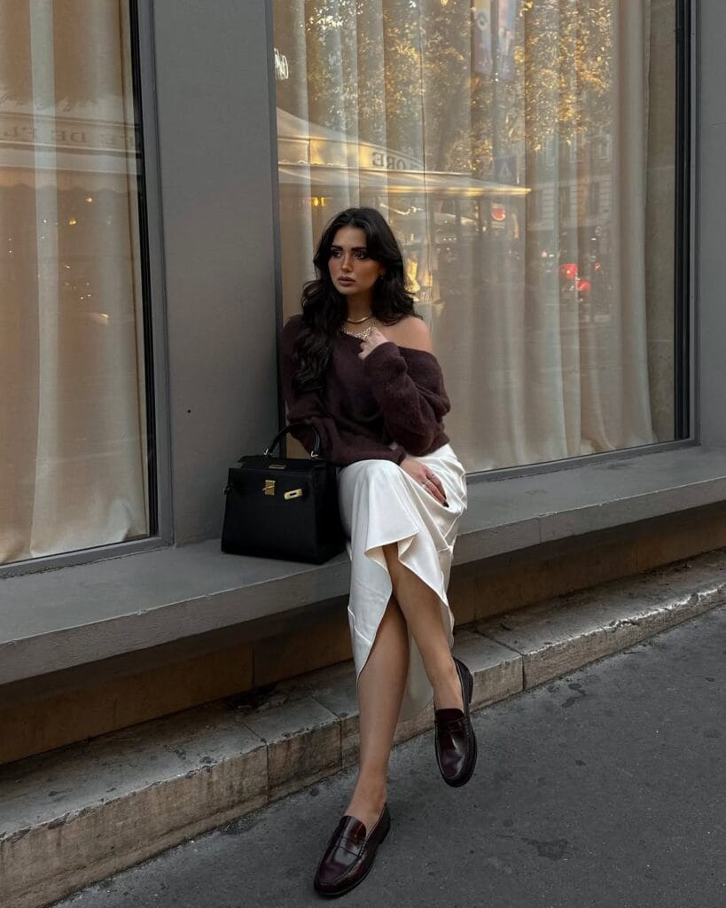Woman in plum off-shoulder sweater, white satin skirt, burgundy loafers, black bag; elegant loafers outfit by window