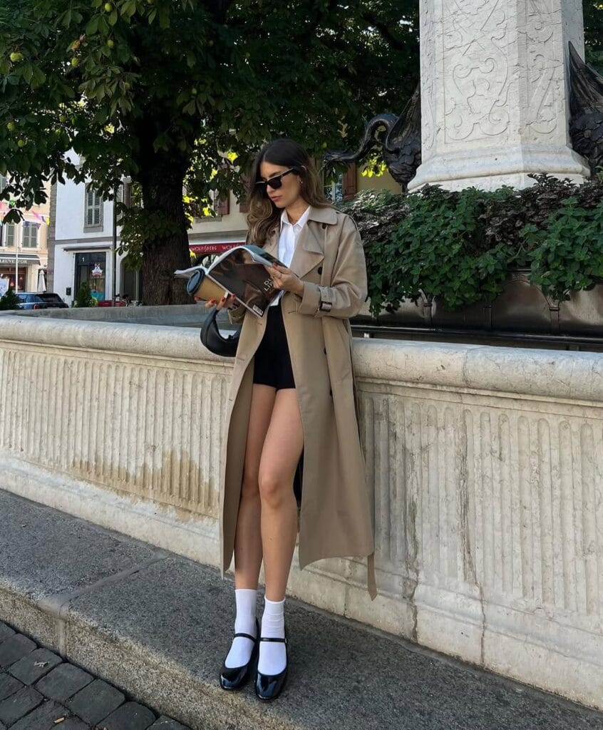 Woman in trench coat, white shirt, black shorts, Mary Janes, and sunglasses reading magazine by fountain