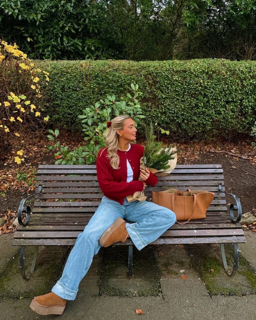 Woman on park bench in red cardigan, jeans, and platform Ugg boots holding flowers; casual winter Ugg outfit.