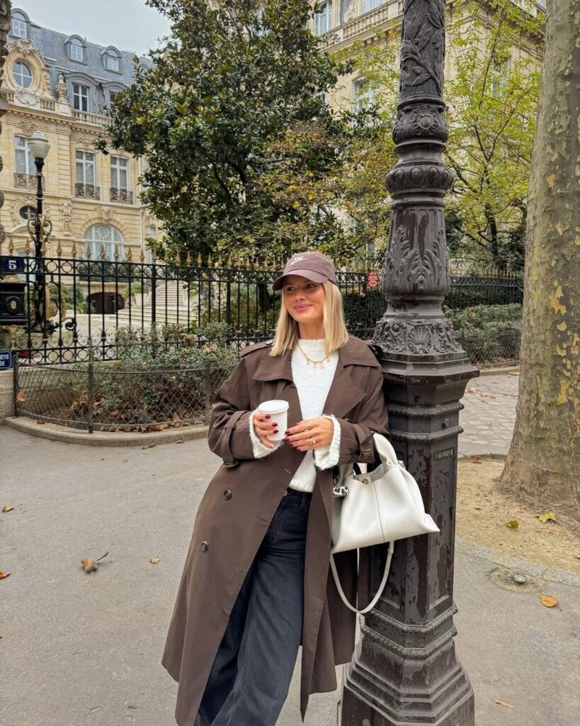 Woman in chocolate trench coat, cream sweater, wide-leg trousers, and cap leaning on lamp post