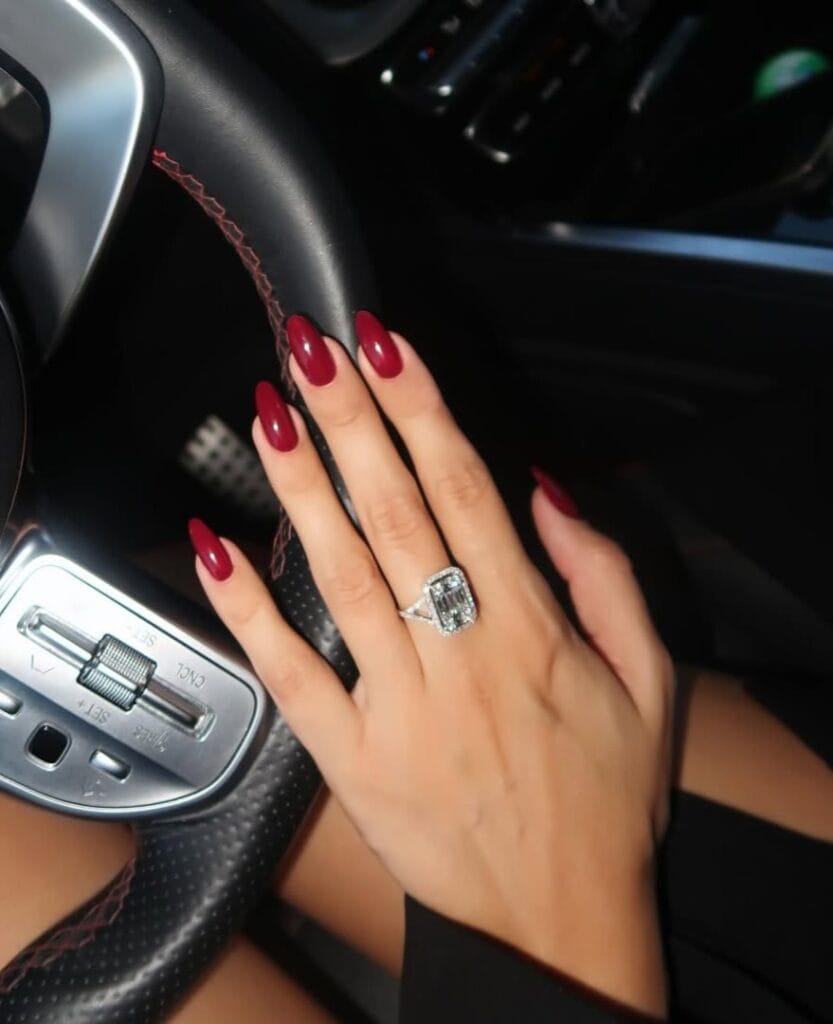 Deep wine red old money nails, almond-shaped, worn with a bold statement ring in car setting.
