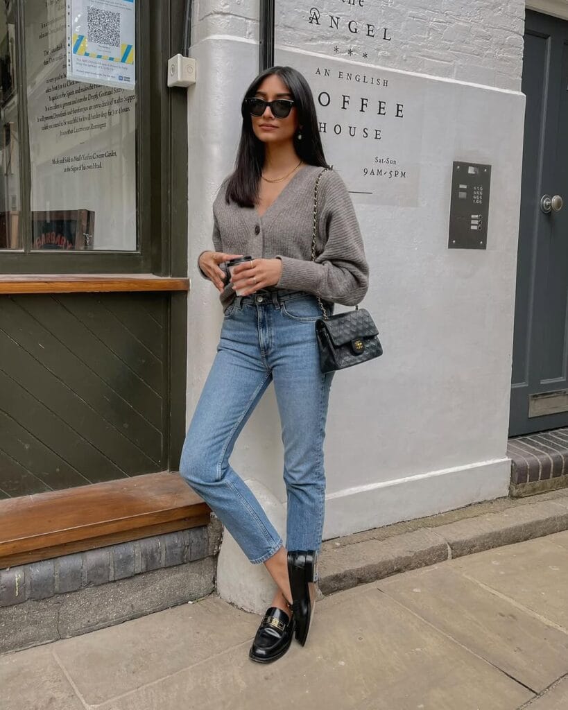 Woman in gray cardigan, blue jeans, black loafers, sunglasses, Chanel bag; casual loafers outfit outside café