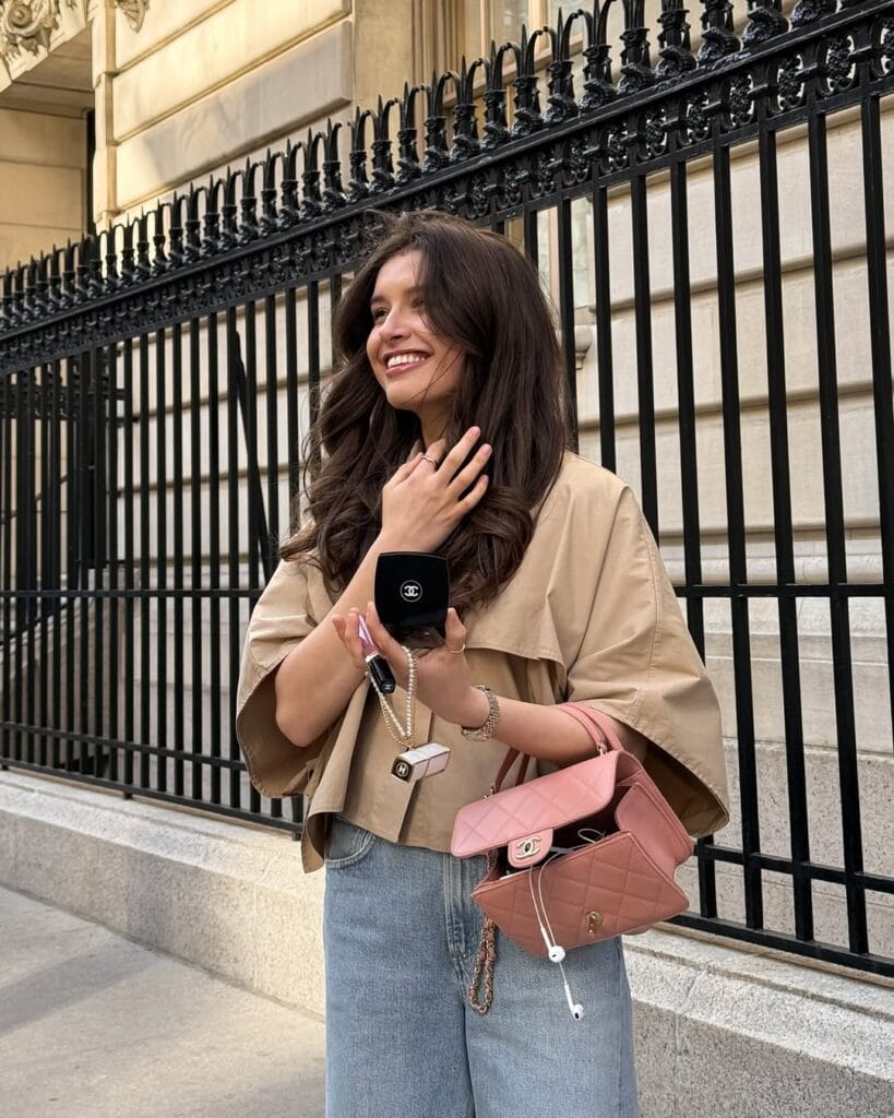Woman in cropped trench coat, light jeans, and pink purse smiling with loose curls in city