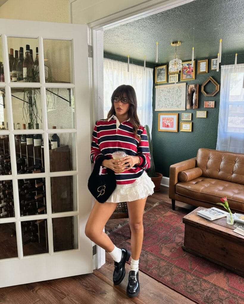 Woman in striped rugby top, ruffled white mini skirt, white socks, black loafers, glasses; preppy loafers outfit indoors
