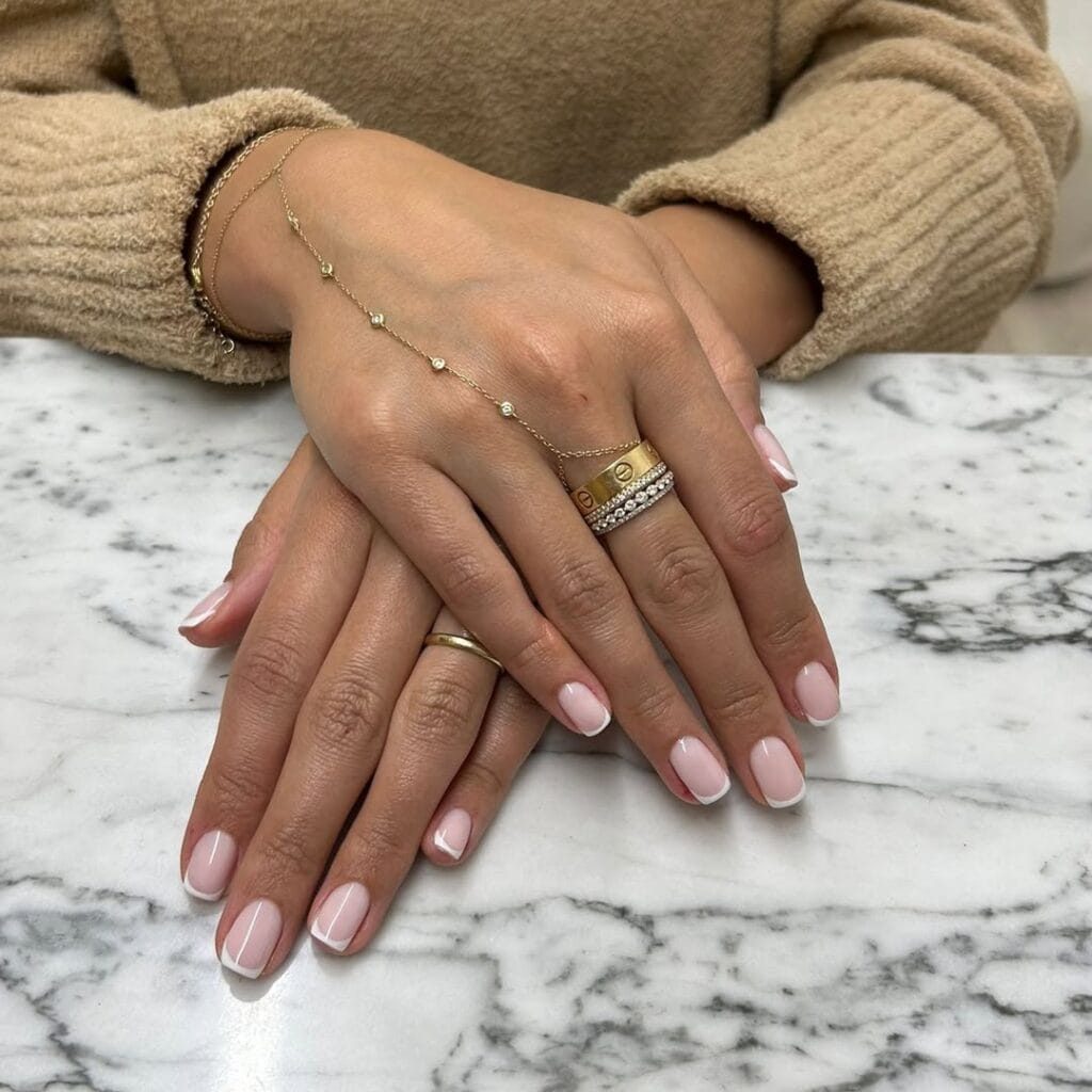 Short French tip old money nails, blush pink base, styled with gold and diamond rings.
