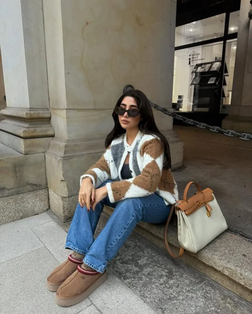 Woman in checkered cardigan, black tank, blue jeans, and Ugg boots with cream bag; chic casual Ugg outfit.
