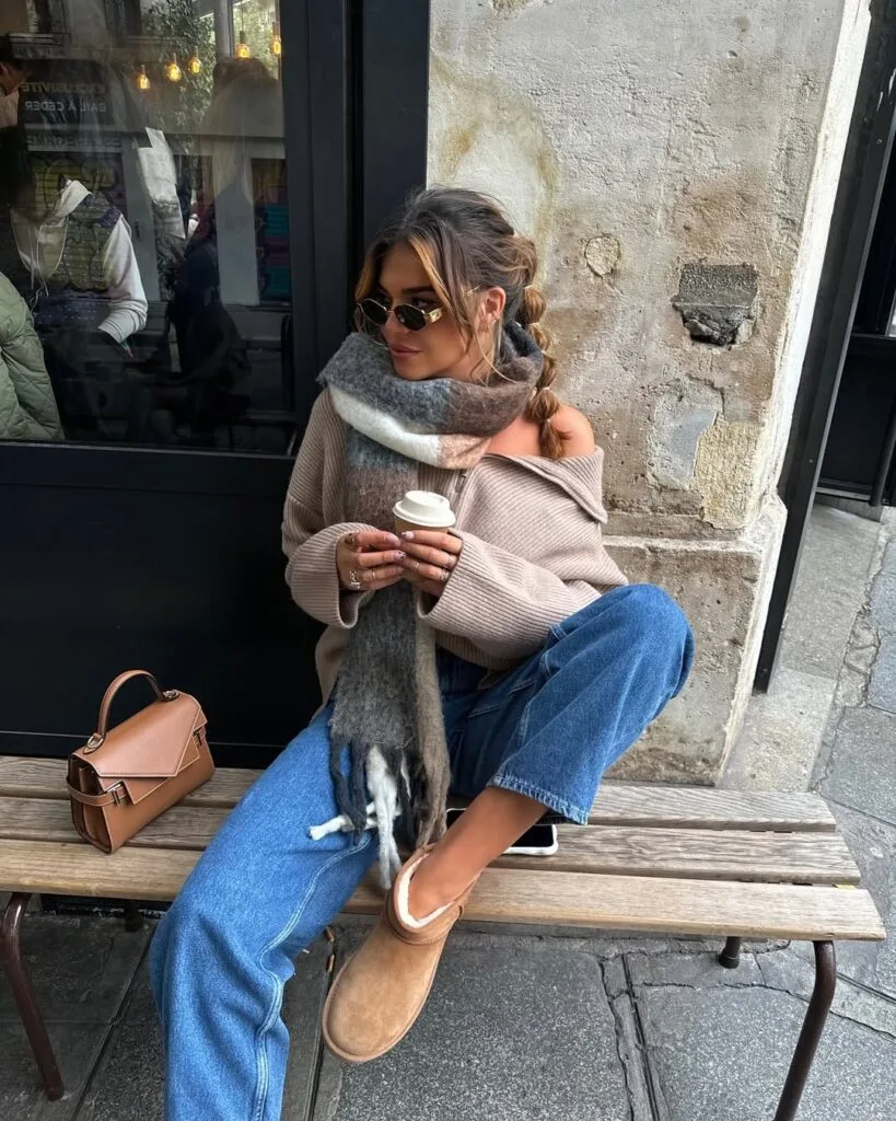 Woman in off-shoulder sweater, oversized scarf, blue jeans, and Ugg boots with tan bag; cozy Ugg outfit.
