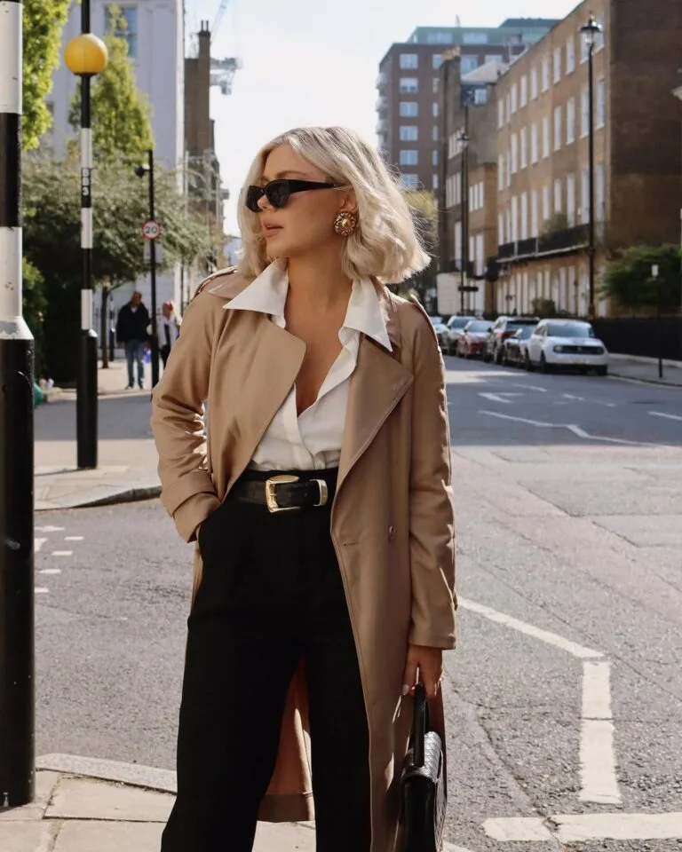 Woman in tan trench coat, white shirt, black pants, gold earrings, and sunglasses walking in city