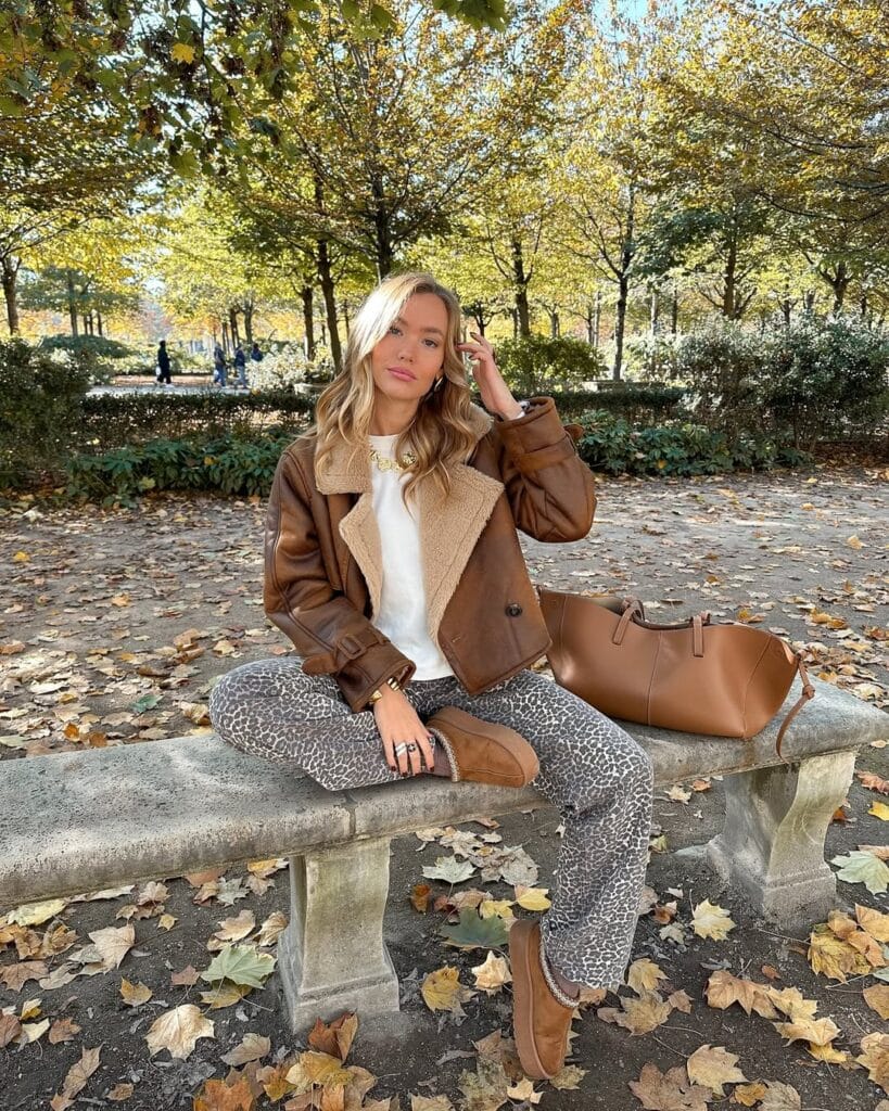 Woman in shearling jacket, animal-print pants, and Ugg boots with tan bag, sitting on bench; trendy Ugg outfit.