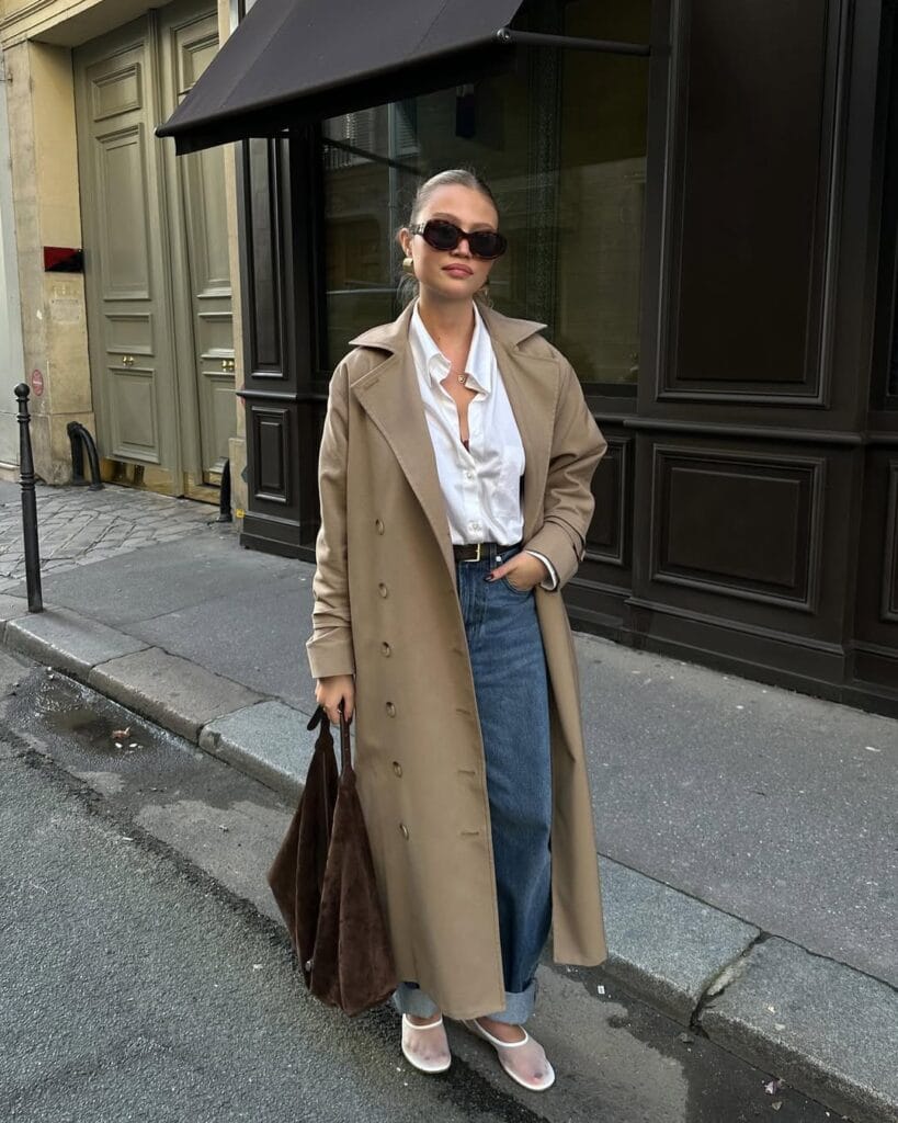 Woman in trench coat, white shirt, blue jeans, mesh ballet flats, and sunglasses holding brown tote