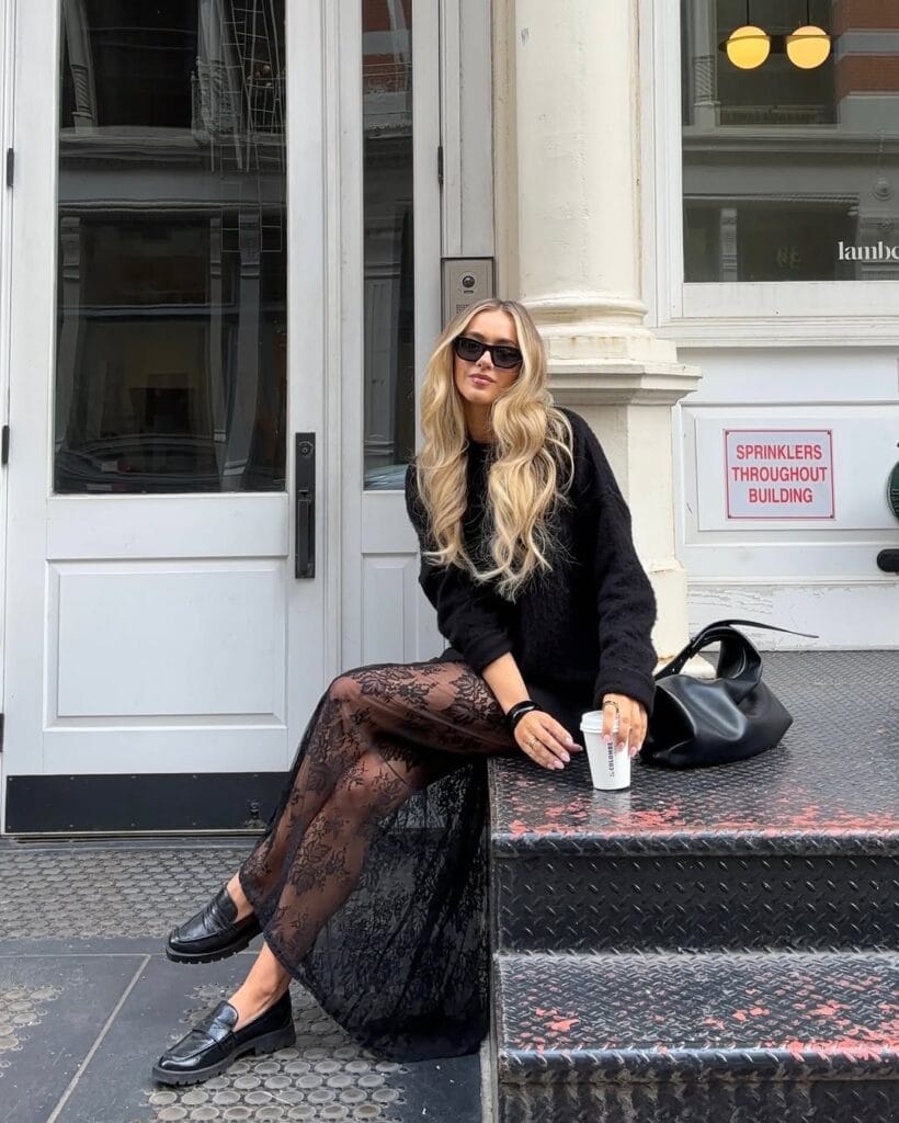 Blonde woman in black sweater, lace maxi skirt, black loafers, sunglasses, coffee cup; lace loafers outfit on city steps
