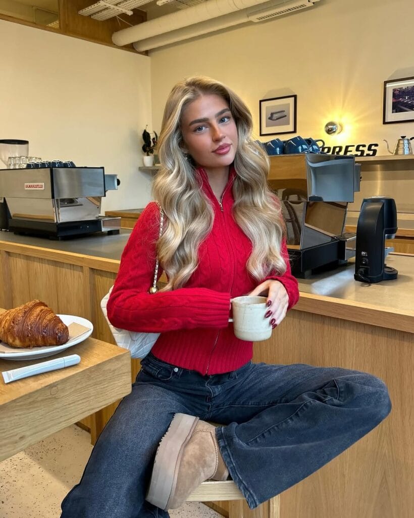 Woman in red cable-knit sweater, dark jeans, and Ugg boots, sitting at café with coffee; stylish casual Ugg outfit.