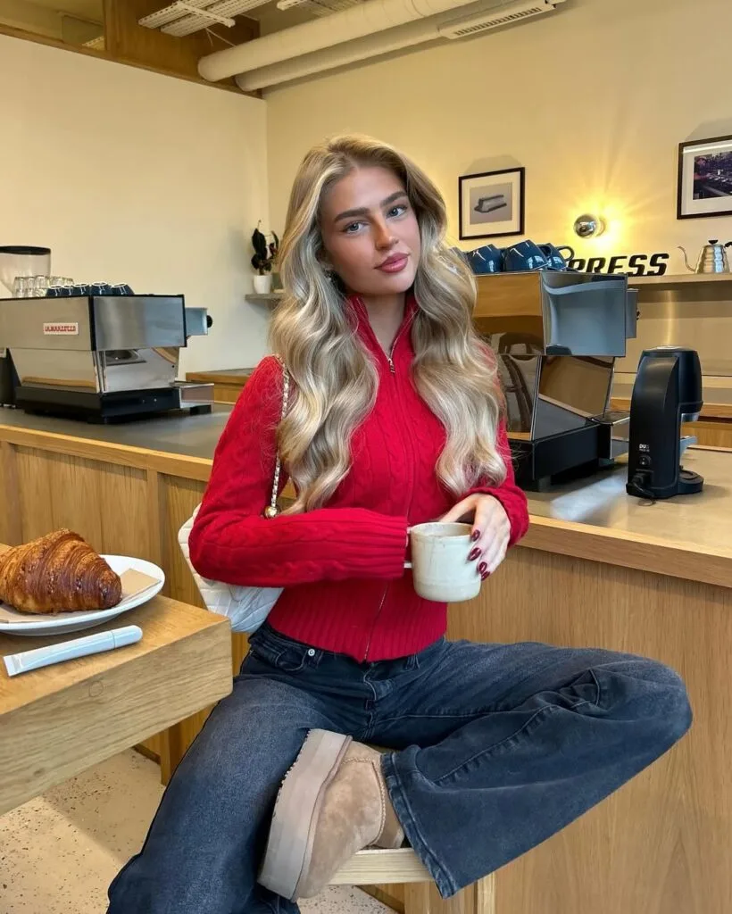 Woman in red cable-knit sweater, dark jeans, and Ugg boots, sitting at café with coffee; stylish casual Ugg outfit.
