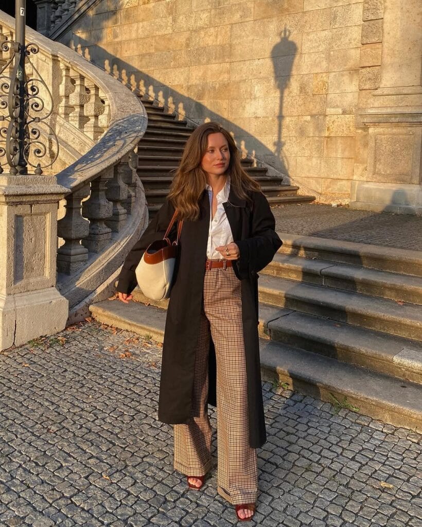 Woman in black trench coat, plaid trousers, white shirt, and sandals standing by stone staircase
