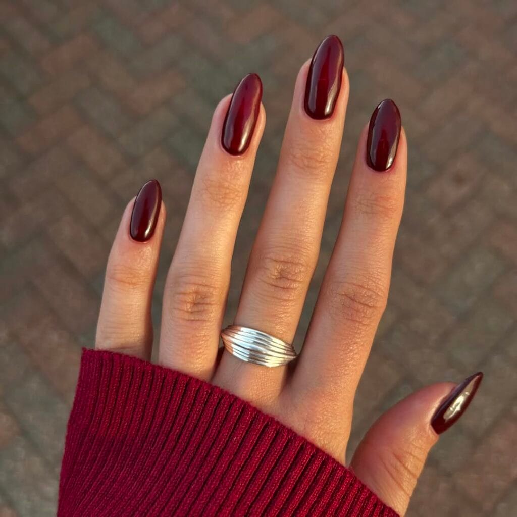 Deep Bordeaux red old money nails, almond-shaped and glossy, styled with silver ring and burgundy sweater.
