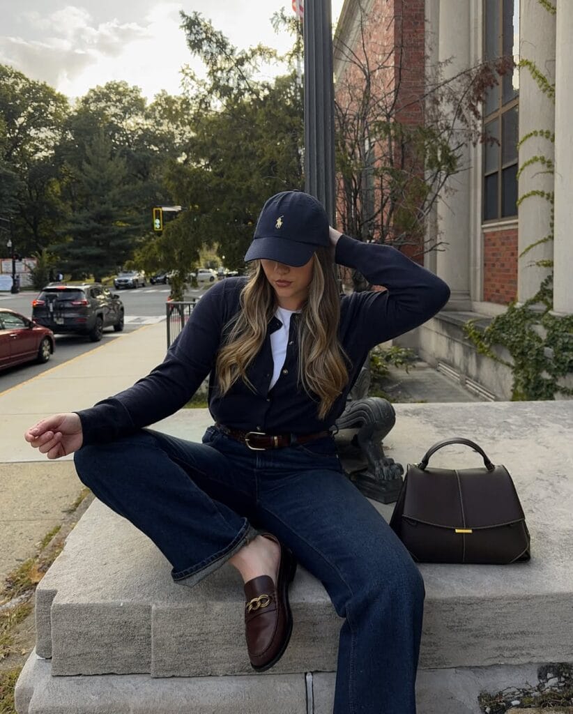 Woman in navy cardigan, white shirt, dark jeans, brown loafers, baseball cap, brown bag; relaxed loafers outfit outdoors