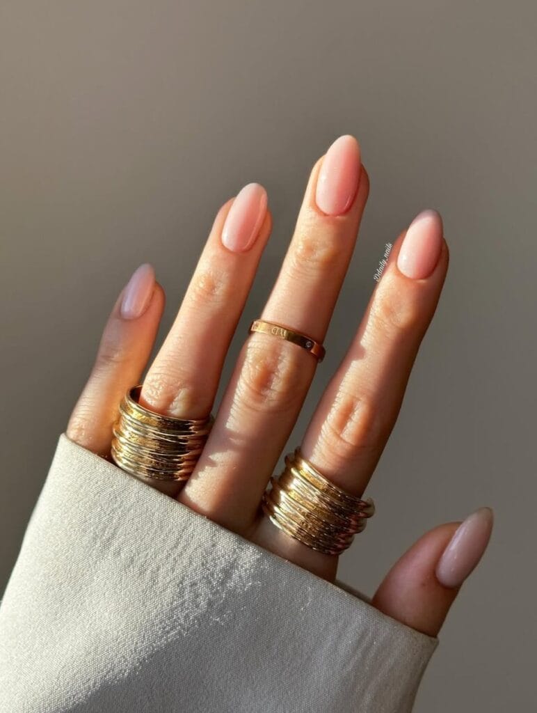 Sheer pink old money nails, almond-shaped, styled with gold stacked rings in natural light.
