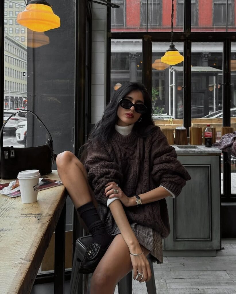Woman in chunky brown sweater, plaid mini skirt, black loafers, black socks, sunglasses; fall loafers outfit in café.