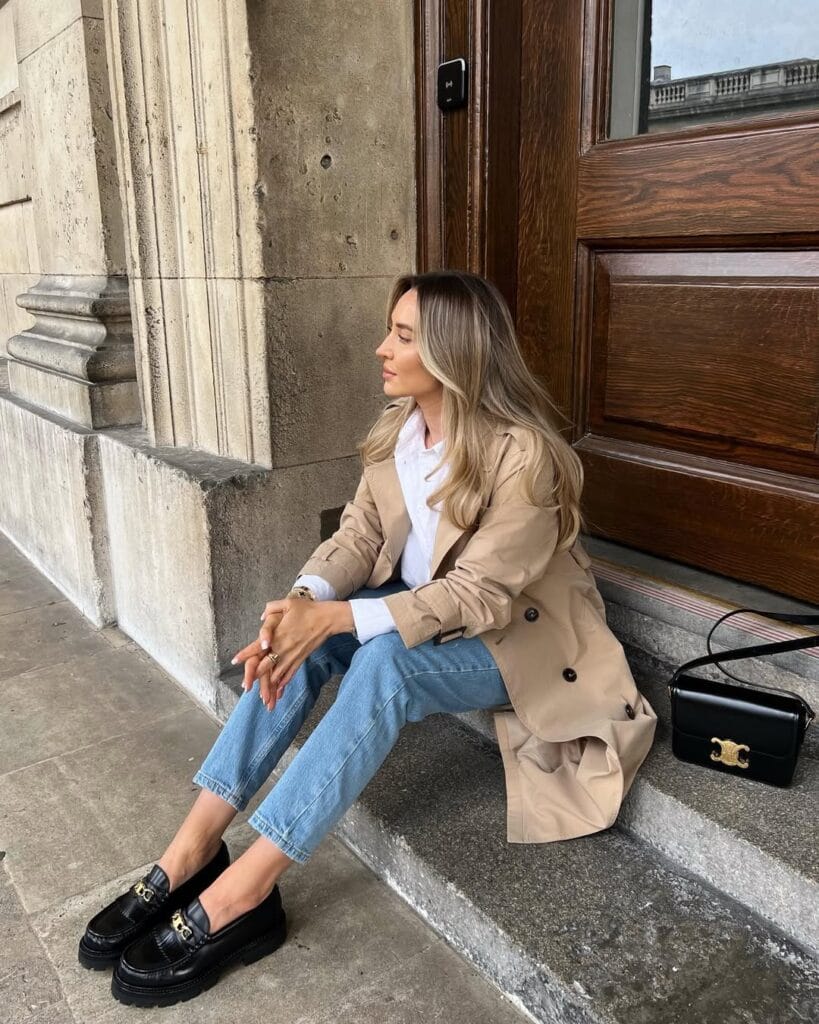 Woman in trench coat, white shirt, blue jeans, and black loafers sitting on steps; loafers outfit, classic city style.