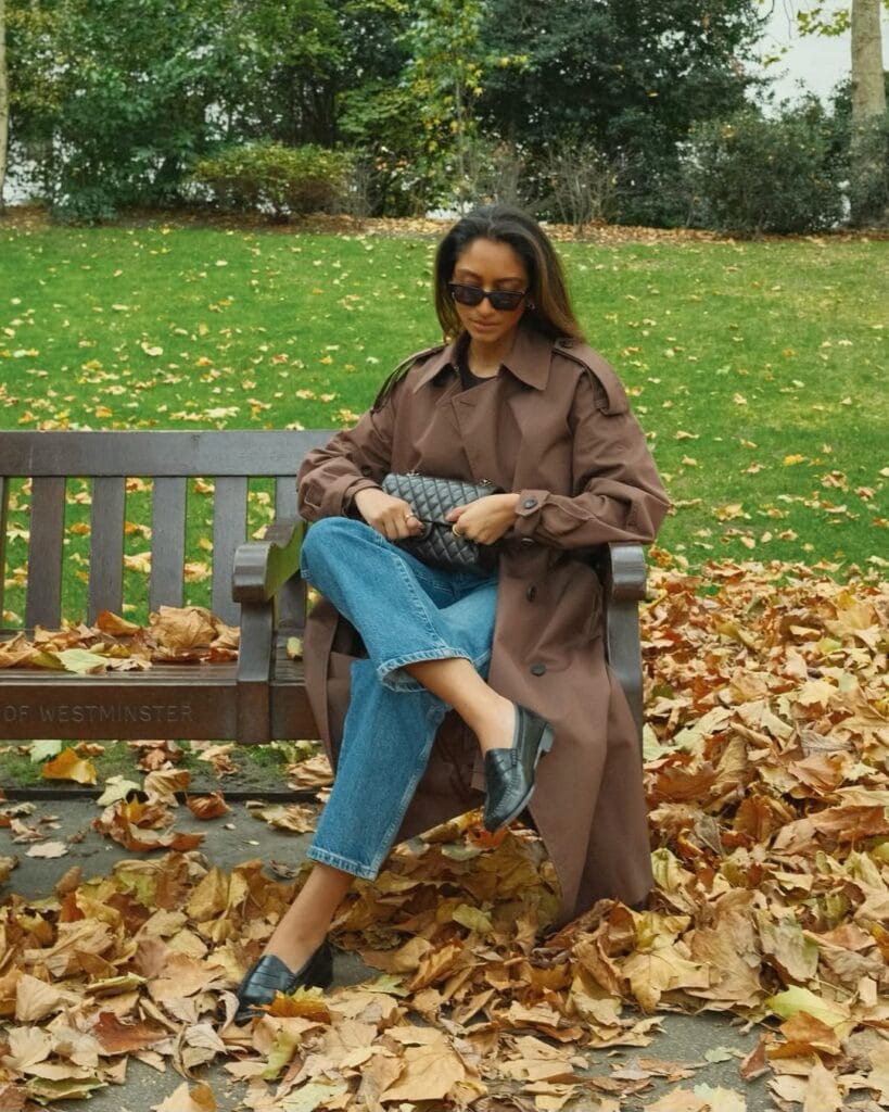 Woman in brown trench coat and jeans sitting on bench with fall leaves; autumn trench outfit